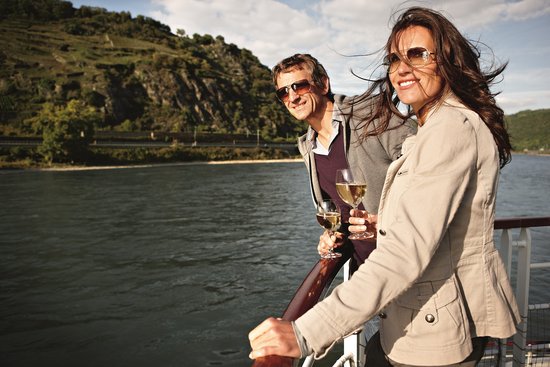 Two people stand on the sun deck of a river cruise ship, enjoying the view of the Rhine with a glass of white wine, surrounded by lush hills and blue skies.