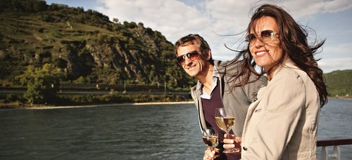 Two people stand on the sun deck of a river cruise ship, enjoying the view of the Rhine with a glass of white wine, surrounded by lush hills and blue skies.