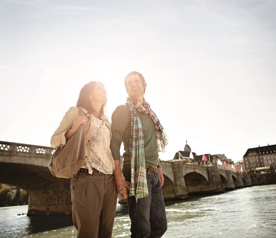 A couple is walking along the banks of the Rhine in Basel in the sunshine. In the background, there is a stone bridge that crosses the river.