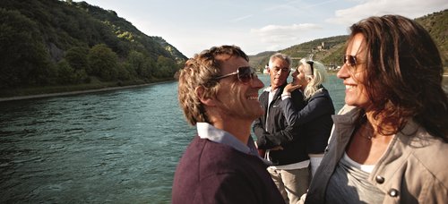 Two couples stand on the sun deck of a river cruise ship, enjoying the view of the Rhine and the surrounding hilly landscape in sunny weather.