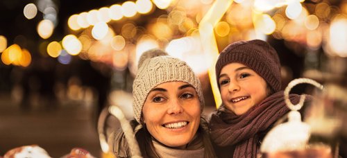 Woman and child with hat and scarf smiling at a Christmas market at night with string lights in the background.