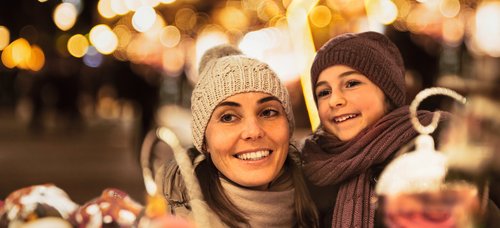 Woman and child with hat and scarf smiling at a Christmas market at night with string lights in the background.
