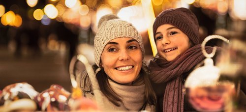 Woman and child with hat and scarf smiling at a Christmas market at night with string lights in the background.