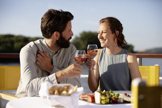 A couple sits outside and clinks glasses of rosé wine, with bread and cheese with grapes on the table.