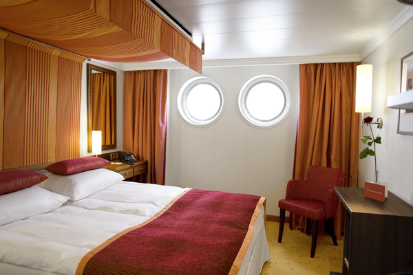 Cabin with two round porthole windows, double bed with red bed runner and pillows, orange curtains, red armchair, and bedside table with lamp and rose.