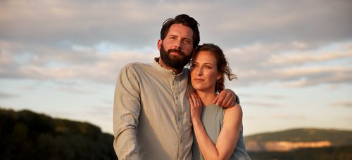 A couple embraces on a boat at dusk with a river and wooded hills in the background.