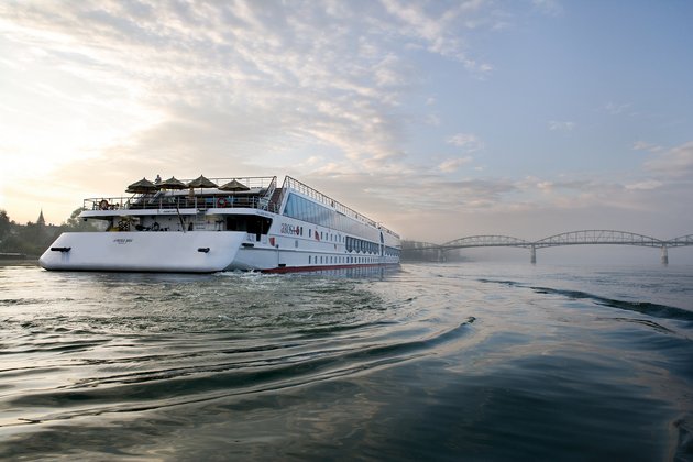 Stern view of the river cruise ship A-ROSA MIA on the Danube at sunrise. In the foreground is the river with gentle waves. In the background is a bridge.
