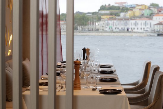 Elegant dining table in the restaurant of the river cruise ship A-ROSA ALVA with set places and a view of the water and a city on the shore.