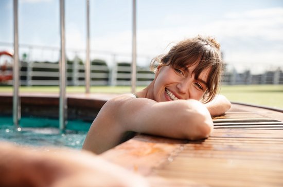 A woman in the pool on the deck of a river cruise ship leans her arms and head relaxed on the edge of the pool and smiles at the person next to her.
