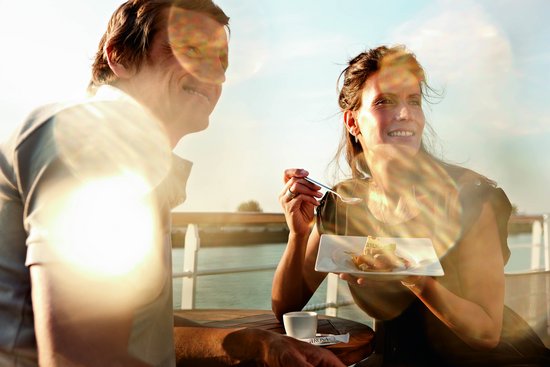 Two people are sitting on the sun deck of a river cruise ship in the sunshine. They are drinking coffee, eating a sweet treat, and looking into the distance. In the background is the river.