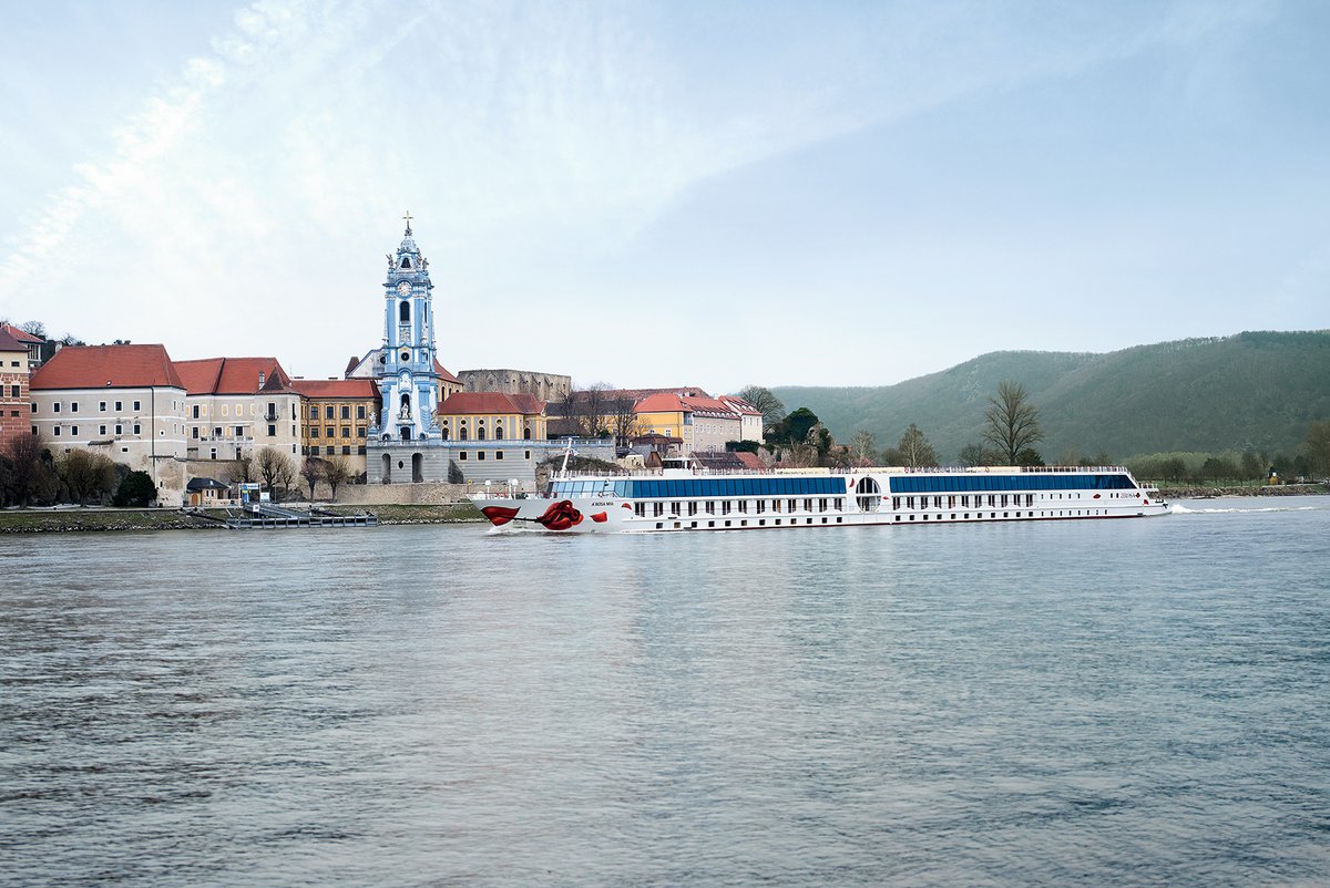 The river cruise ship A-ROSA MIA passes by Dürnstein Abbey on the Danube. In the foreground, the river; in the background, a hilly landscape covered with vegetation under a clear sky.