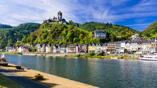 Blick auf die malerische Stadt Cochem mit der Reichsburg auf einem Hügel, umgeben von grünen Weinbergen und dem Fluss Mosel im Vordergrund.