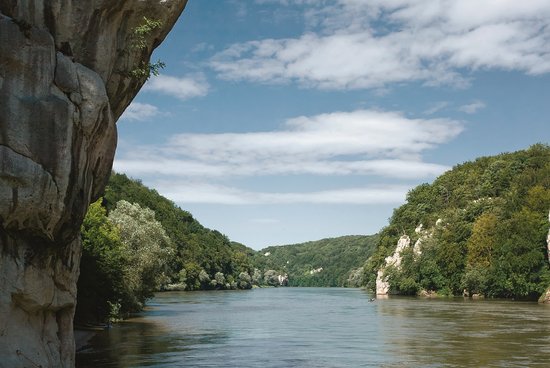View of the Danube on the cataract section with a rock face on the left in the foreground and canoeists on the river, surrounded by vegetated hills under a cloudy sky.