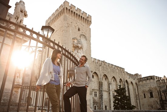 A couple stands in the old town of Avignon in front of a fence at a historic building. In the background, the sun is setting.