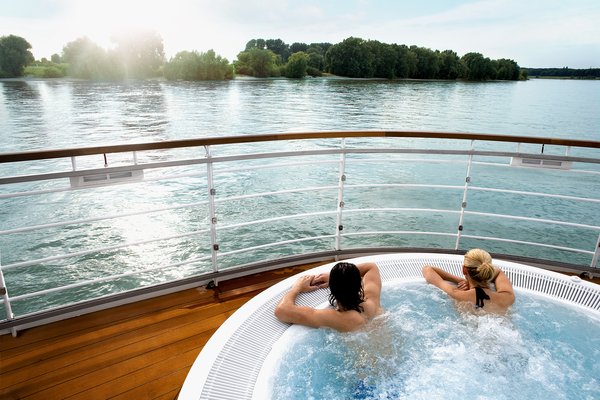 Two people relax in the afternoon in the hot tub on deck with a view of the calm river and green riverbank landscape.