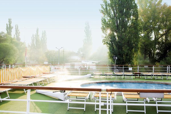 The sun deck of the river cruise ship A-ROSA RIVA with deck chairs and a pool in the morning mist. In the background is the port of Esztergom on the Danube.