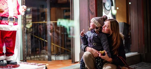 Mother and child are looking at a shop window with a Santa Claus figure together, the child is holding a lollipop and the mother a piece of pastry.