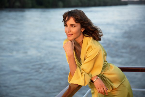 An elegantly dressed woman leans against the railing of a river cruise ship and watches the sunset over the river, while she relaxes and enjoys the view.