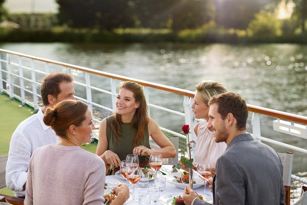 Yvonne Catterfeld and four other people are sitting at a set table on a sun deck by the water, enjoying a meal at sunset.