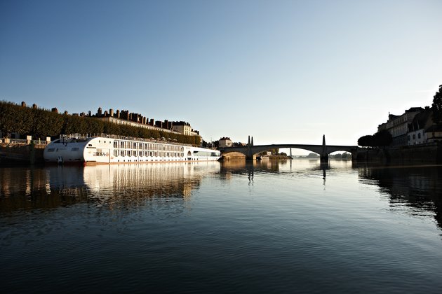 The A-ROSA STELLA is moored on the bank in a city on the Rhône at sunrise, with a bridge and buildings in the background.