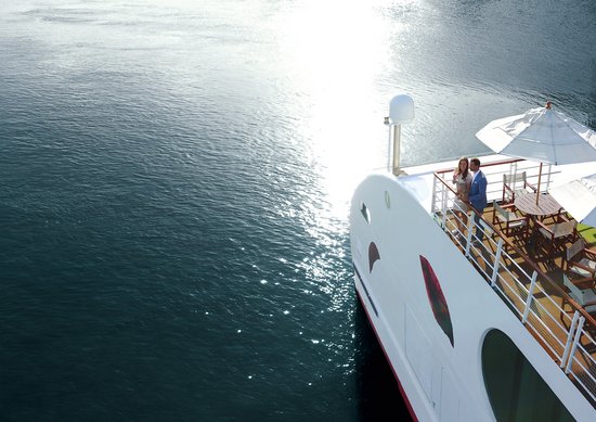 Couple standing on the sun deck of an A-ROSA ship with sun umbrellas, looking at the calm water with sun reflections.