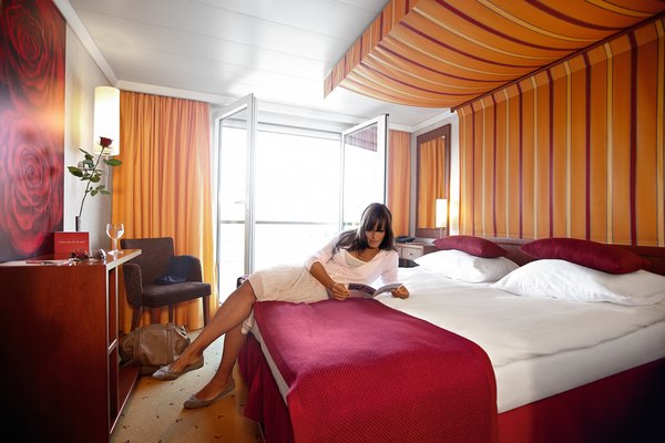 A cabin of category C and D on board the A-ROSA LUNA and A-ROSA STELLA. Furnished in warm colors with a double bed and a French balcony. A woman is lying on the bed reading a newspaper.