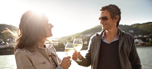 Two people are standing on the sun deck of a river cruise ship, clinking glasses of white wine, surrounded by green hills with houses and the Rhine in the background.