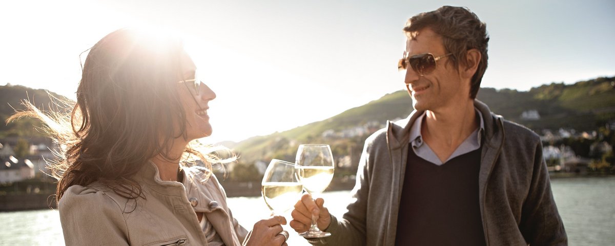 Two people are standing on the sun deck of a river cruise ship, clinking glasses of white wine, surrounded by green hills with houses and the Rhine in the background.