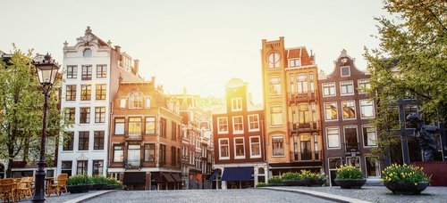 View of Amsterdams historic buildings at sunset. In the foreground is a cobblestone path surrounded by trees, flower pots, a lantern, and seating under a clear sky.