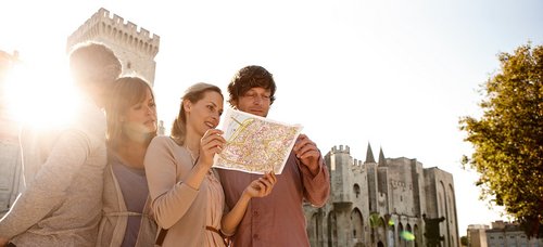 Two couples stand in Avignon in front of a historic building and look at a city map. In the background, the sun is setting.
