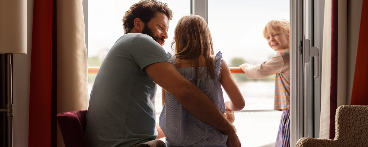 A father is sitting with his child in a family cabin on a river cruise ship in front of the balcony. A second child is standing on the balcony.