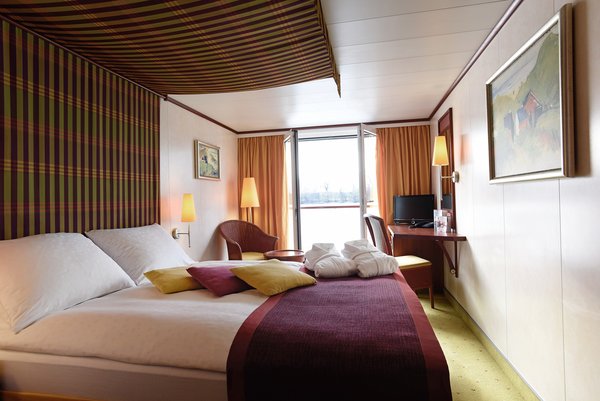A category C cabin on a Danube ship with a double bed and an open French balcony, as well as seating options and a desk with a television.