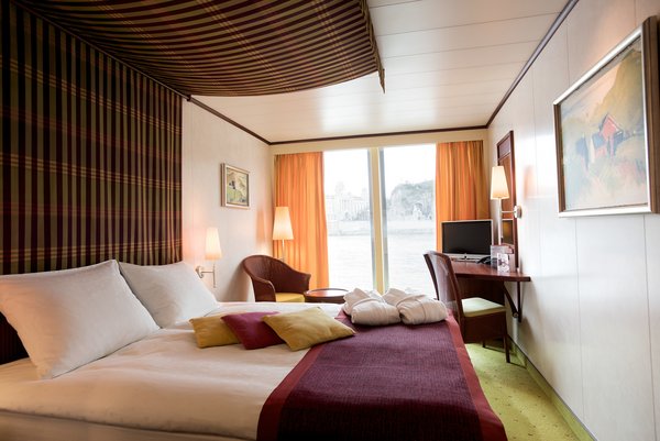 A category B cabin on a Danube ship with a double bed and panoramic window, as well as seating options and a desk with a television.
