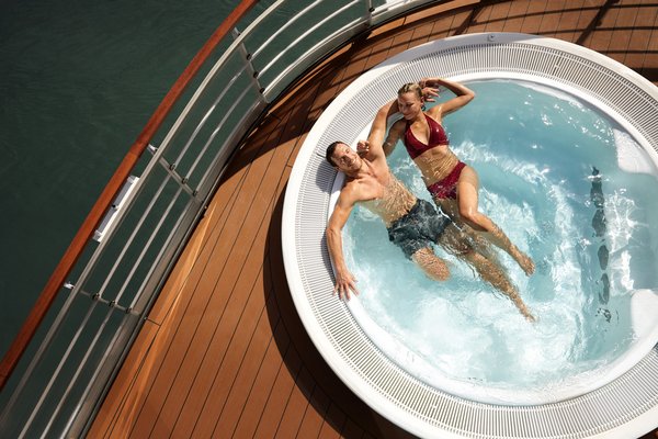 Couple relaxing in a round hot tub on a wooden deck with water and metal railing in the background.