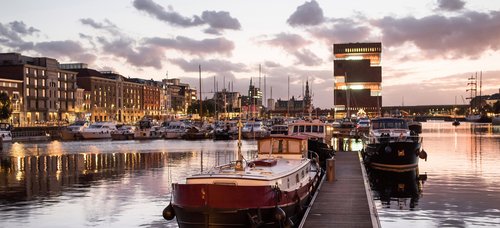 Port in Antwerp at dusk. In the port are historical boats and yachts. In the background, historical and modern buildings.