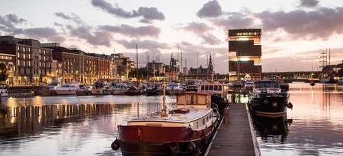 Port in Antwerp at dusk. In the port are historical boats and yachts. In the background, historical and modern buildings.