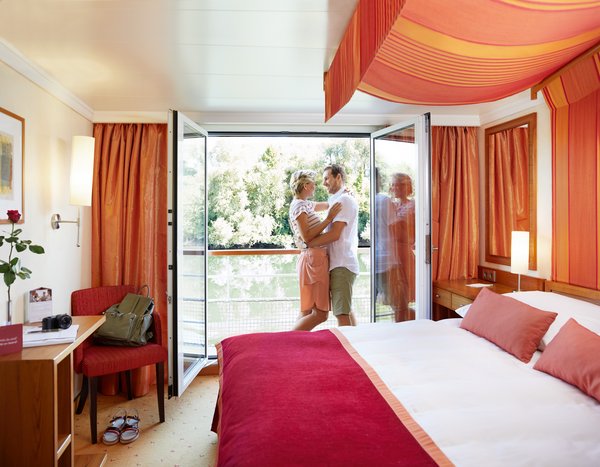 A couple stands on the balcony of a cabin overlooking green trees, in the room a large bed with a red throw and orange curtains.