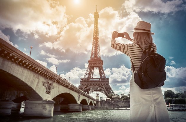 A tourist with a straw hat and backpack photographs the Eiffel Tower under a sunny sky from the Seine bridge in Paris.