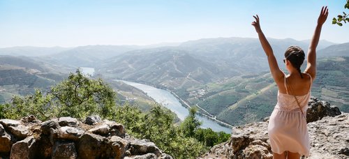 Woman in a white dress stands on rocks and raises her arms over a river and green hills in the valley.