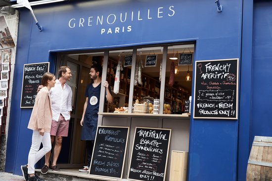 Blue shop with the sign Grenouilles Paris, two customers talking to a seller at the open window, boards with French dishes and drinks.