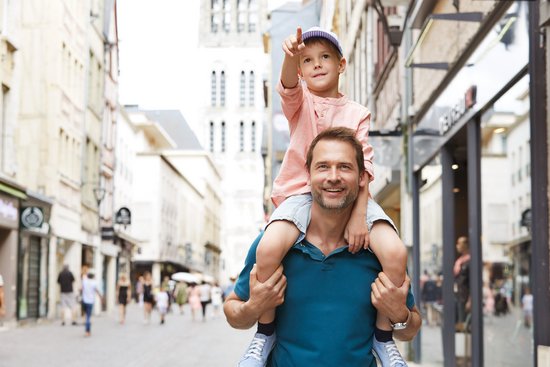 Father carries son on his shoulders in a busy shopping street with historic buildings in the background.