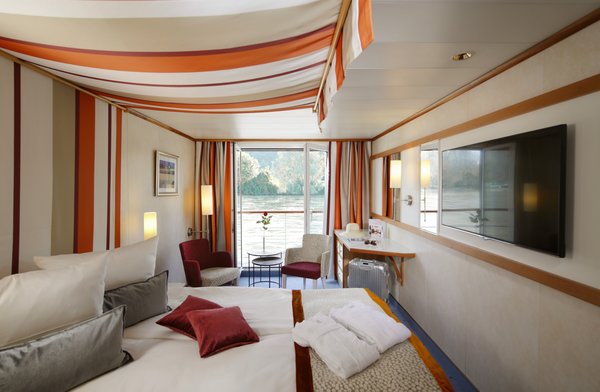 A category C cabin on the A-ROSA DONNA with a double bed and an open French balcony, as well as seating options, a desk, and a flat-screen TV, with luggage and bathrobes in the cabin.