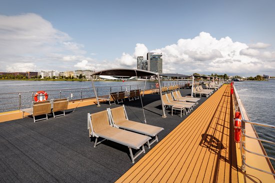 The sun deck with numerous deck chairs and parasols on a sunny day with a slightly cloudy sky. In the background, Amsterdam.
