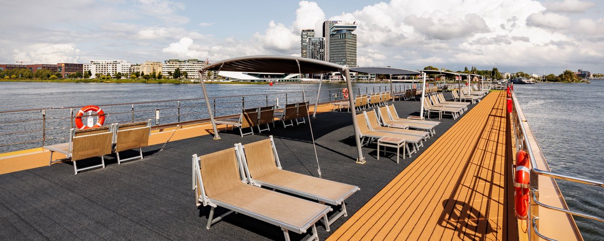 The sun deck with numerous deck chairs and parasols on a sunny day with a slightly cloudy sky. In the background, Amsterdam.