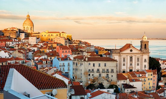 Panorama of Lisbon with red tiled roofs, historic buildings, and a large church with a dome in the warm evening light.