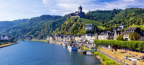 Blick auf die malerische Stadt Cochem mit der Reichsburg auf einem Hügel, umgeben von grünen Weinbergen und dem Fluss Mosel im Vordergrund.