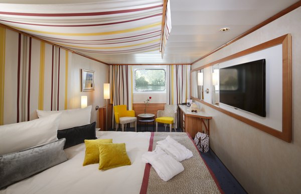 A category A cabin on the A-ROSA DONNA with a double bed and window, seating options, a desk and flat-screen TV, with luggage and bathrobes in the cabin.