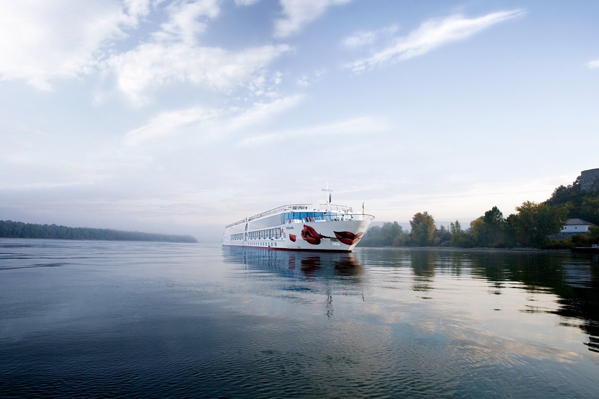 The river cruise ship A-ROSA MIA on the Danube. The calm river with trees on the shore surrounds the ship with the distinctive kiss mark and the rose on the bow.