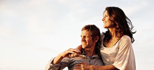 A woman sits on the railing of a river cruise ship and places her arms on the man next to her, who is leaning against the railing and holding her. They enjoy the view of the Rhine in the sunshine.