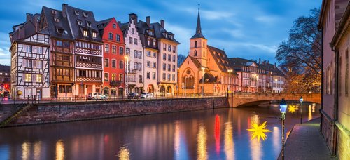 Evening view of the historic half-timbered houses and a church on the riverbank in Strasbourg with illuminated street lamps and reflections in the water.
